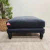 Isla Medium Square Footstool In Armour Smart Velvet 1 A dark blue ottoman with turned legs sits on a light-colored rug. A triangular warning tag is attached to the side. A potted plant is visible in the background.