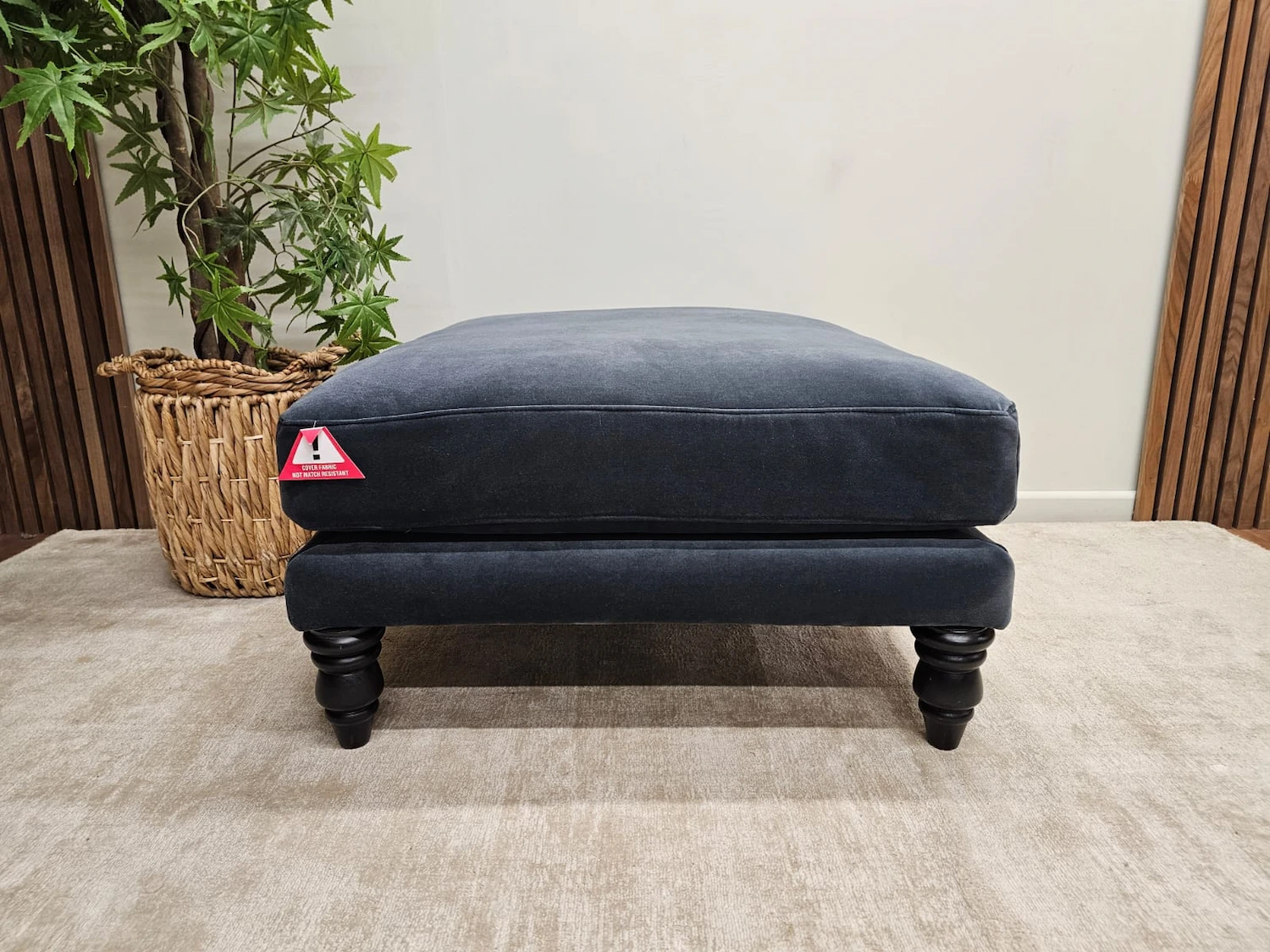 A dark blue ottoman with turned legs sits on a light-colored rug. A triangular warning tag is attached to the side. A potted plant is visible in the background.